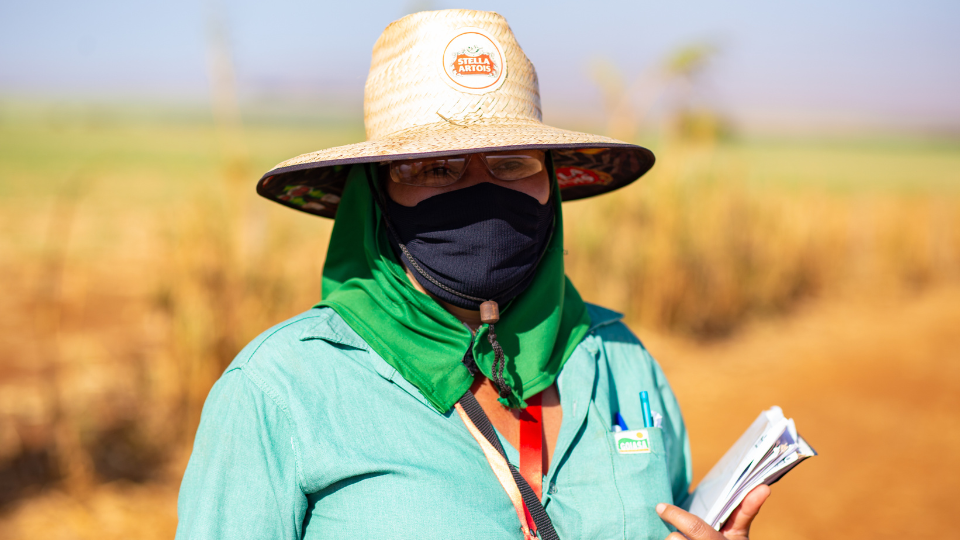 A woman wearing protective gear (a straw hat and mask over her mouth) and holding clipboard outside on a Fair Trade Certified Sugar Farm